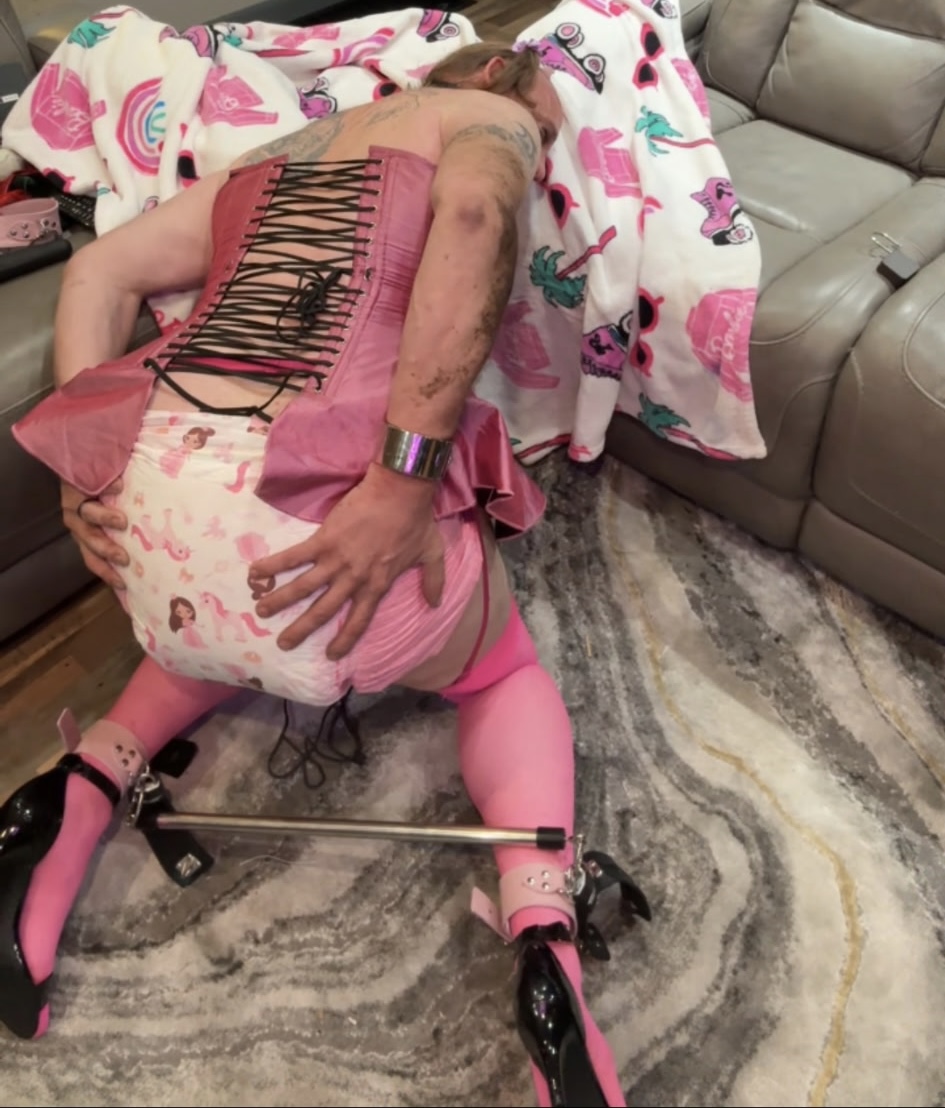 A person in pink stockings and a laced corset lies on the floor beside a sofa, one hand resting on a printed diaper.
