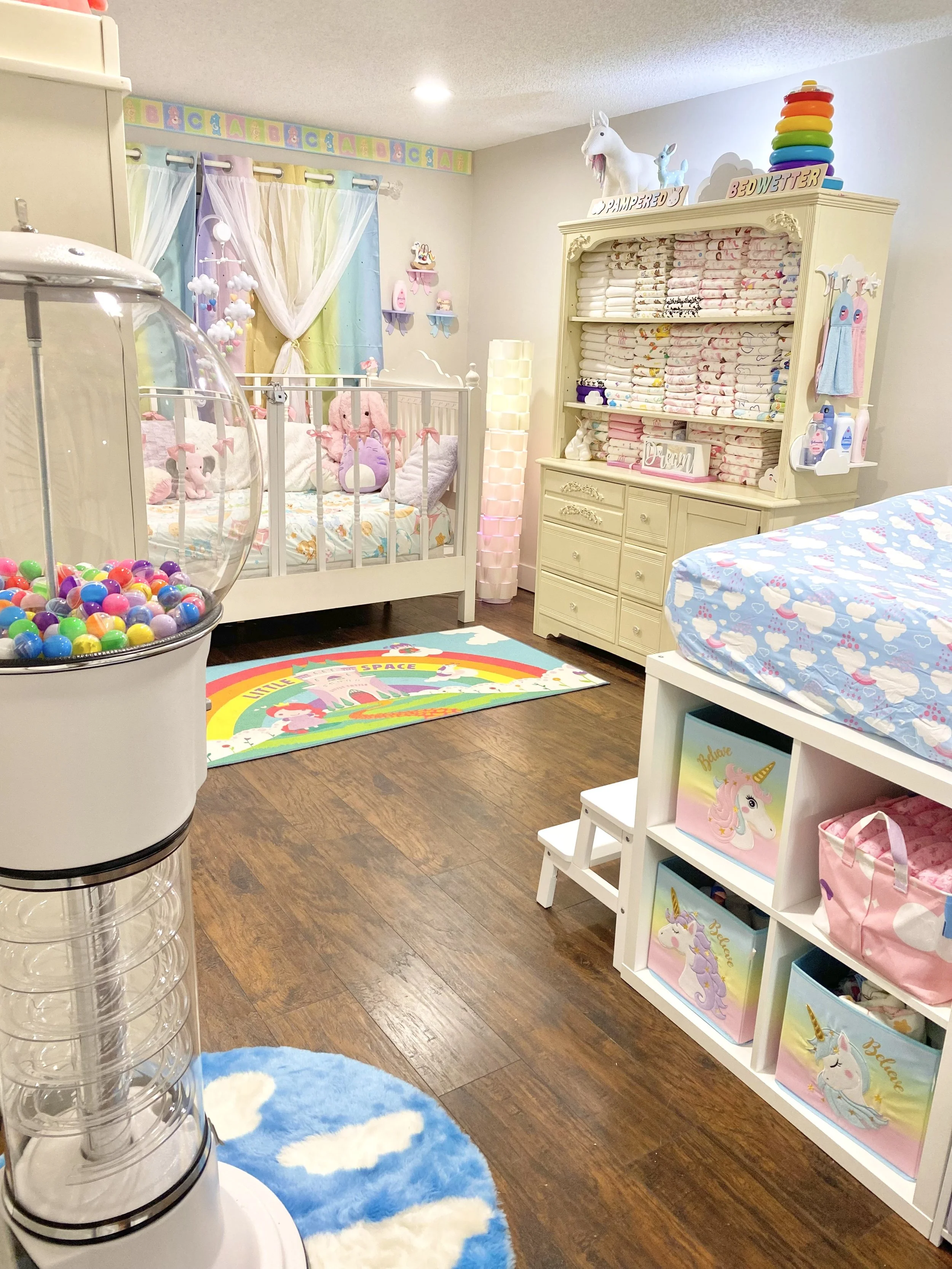 A pastel nursery with a crib, diaper-filled dresser, unicorn storage bins, and a gumball-style dispenser.