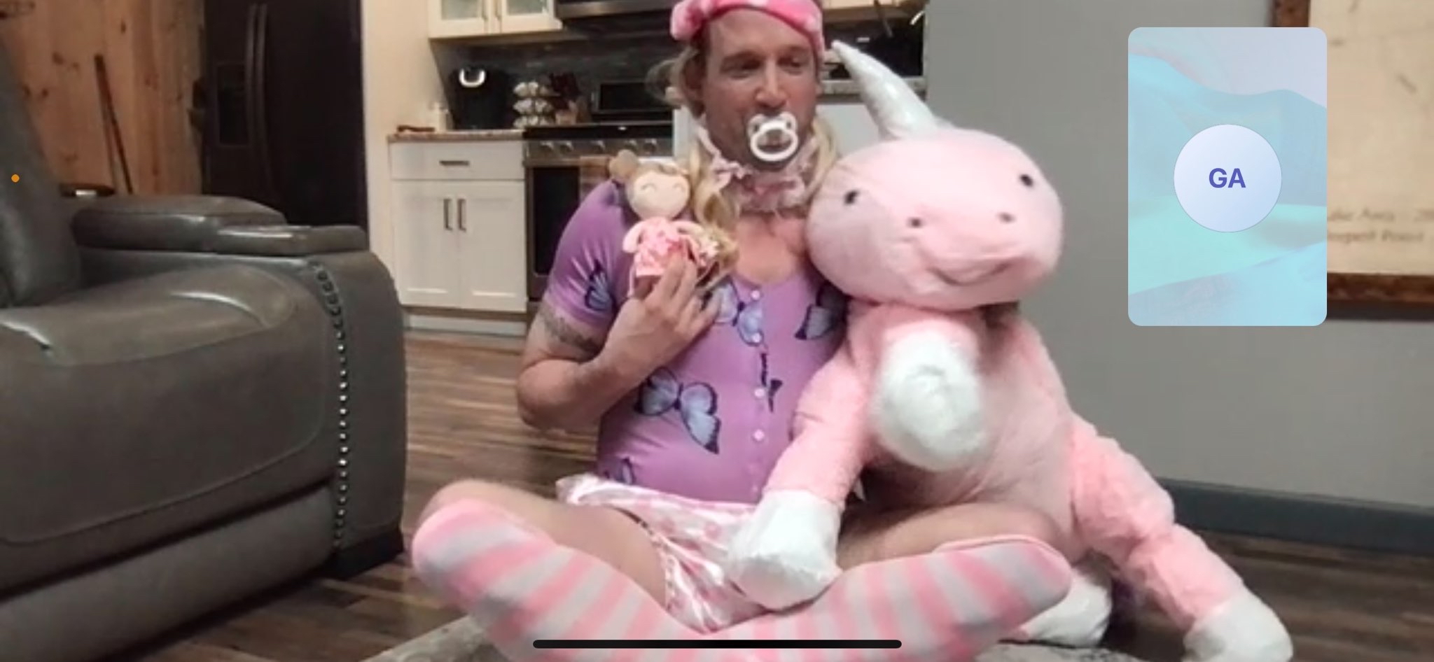 A person sitting on the floor holds a doll beside a large pink plush animal in a room with a kitchen behind them.