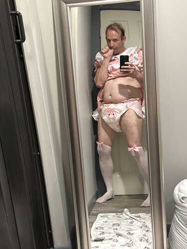 A person stands in a doorway mirror wearing a pink frilly top, diaper, and white stockings while taking a selfie.