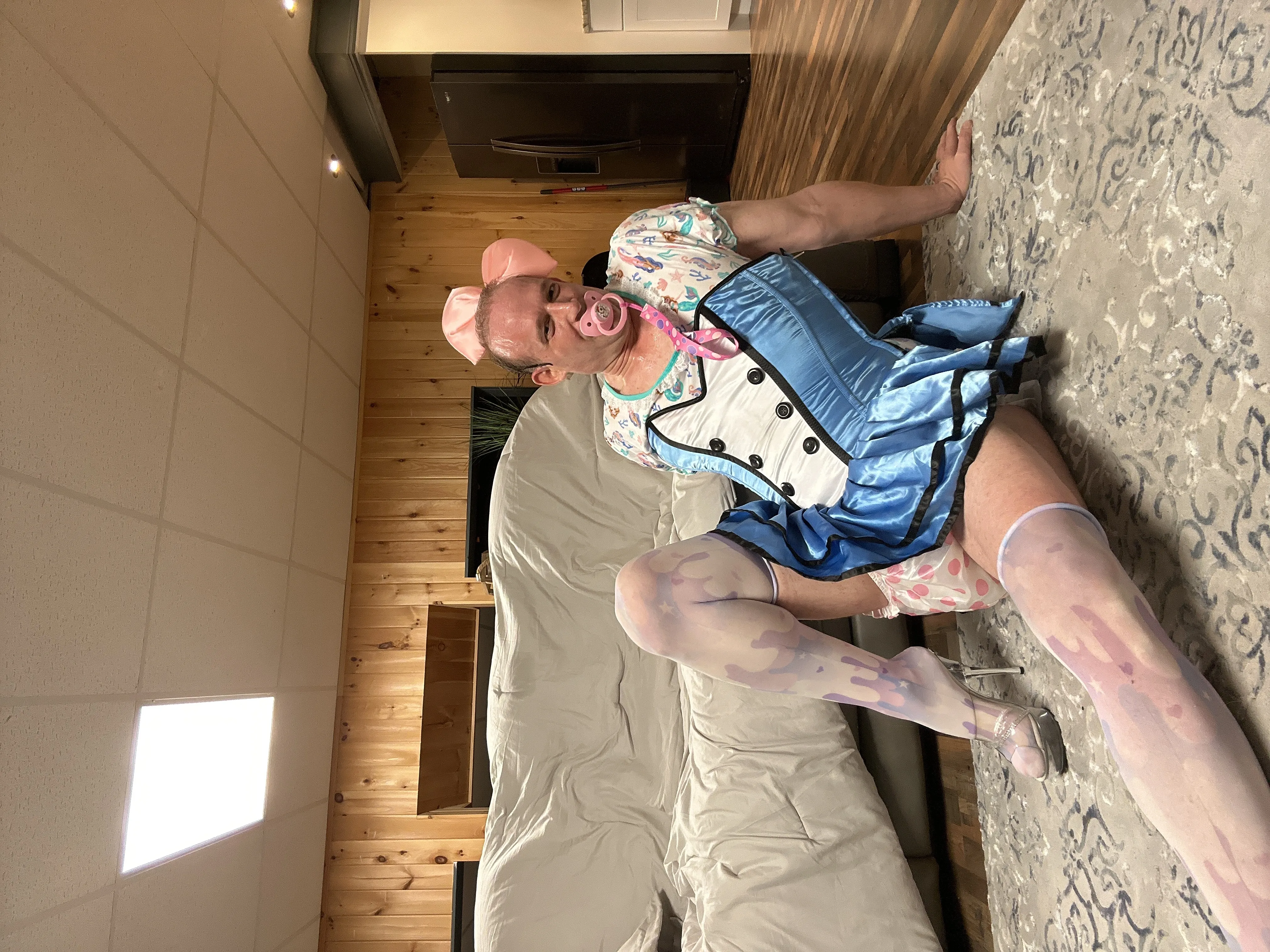 A person in a blue costume sits sideways on a sofa in a wood-paneled room with a pink bow and pacifier.