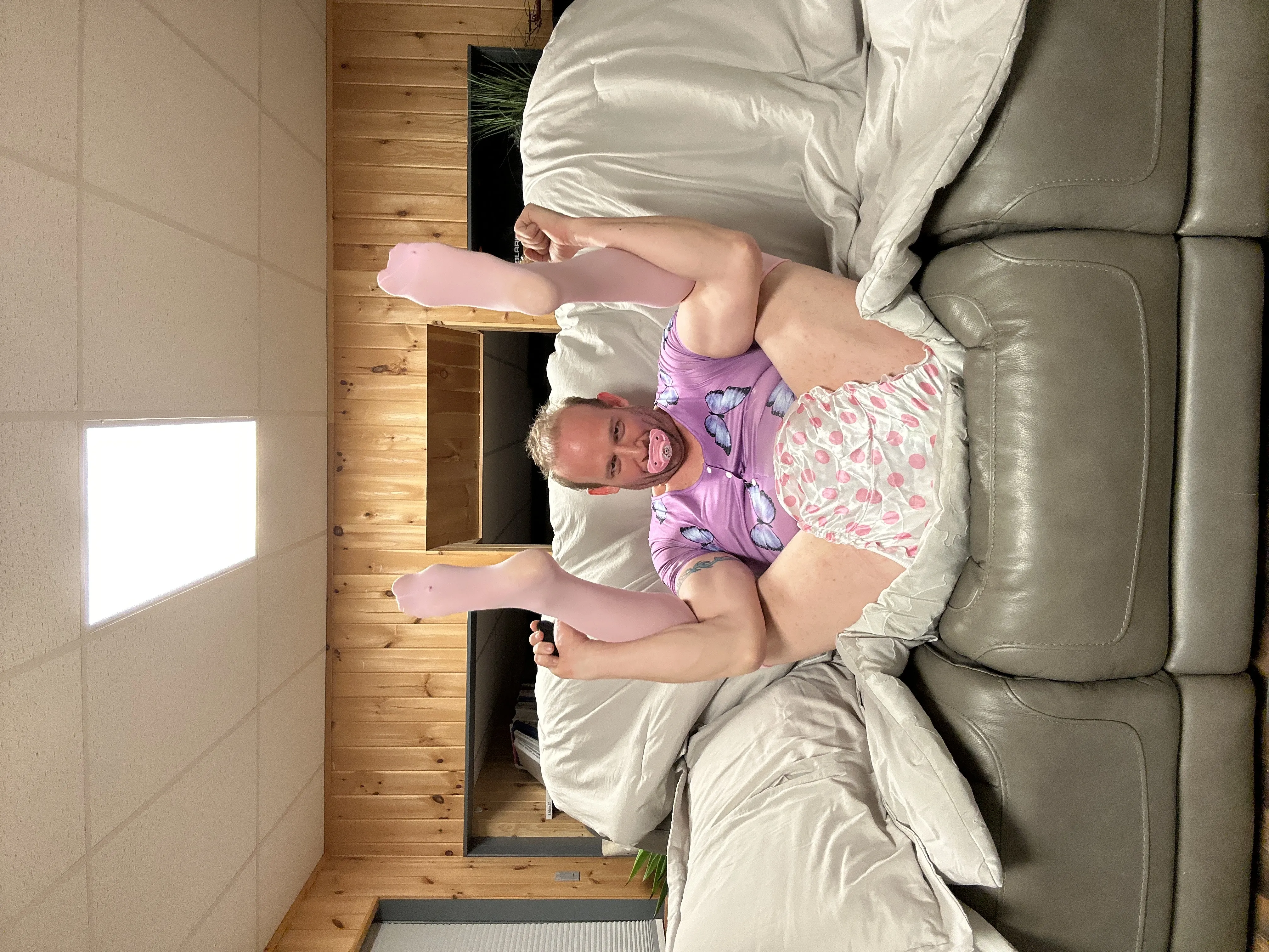 A sideways view of a person on a bed with one leg raised, wearing a purple top and diaper in a wood-paneled room.