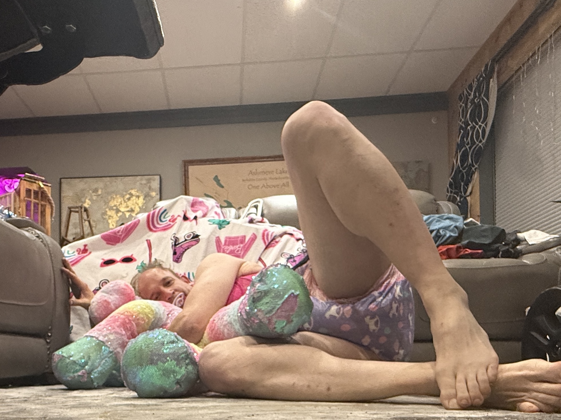 A person reclines on the floor beside a sofa, resting against a pastel plush toy in a cluttered living room.
