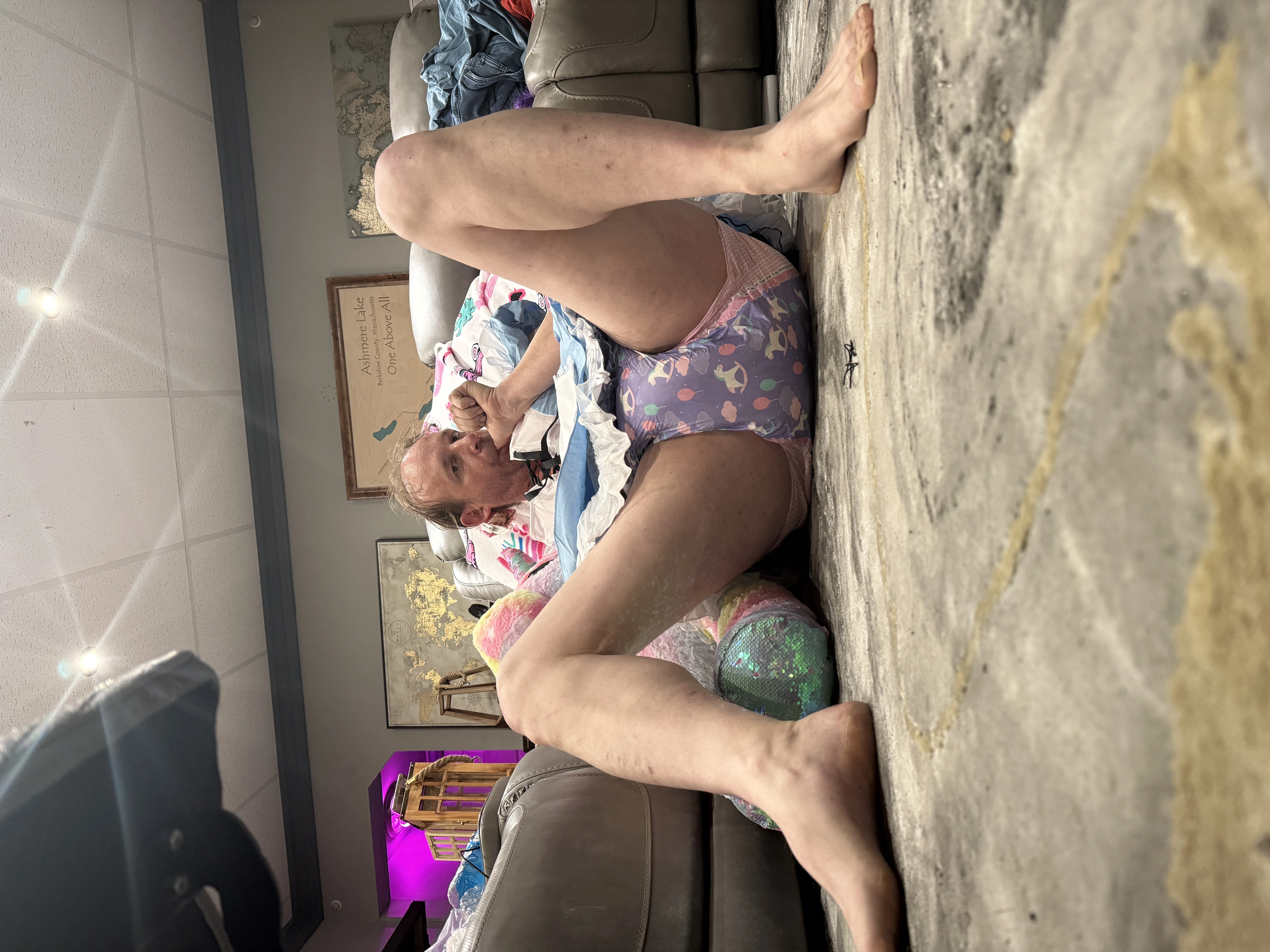 A sideways photo shows a person seated on the floor with bent legs in a cluttered living room.