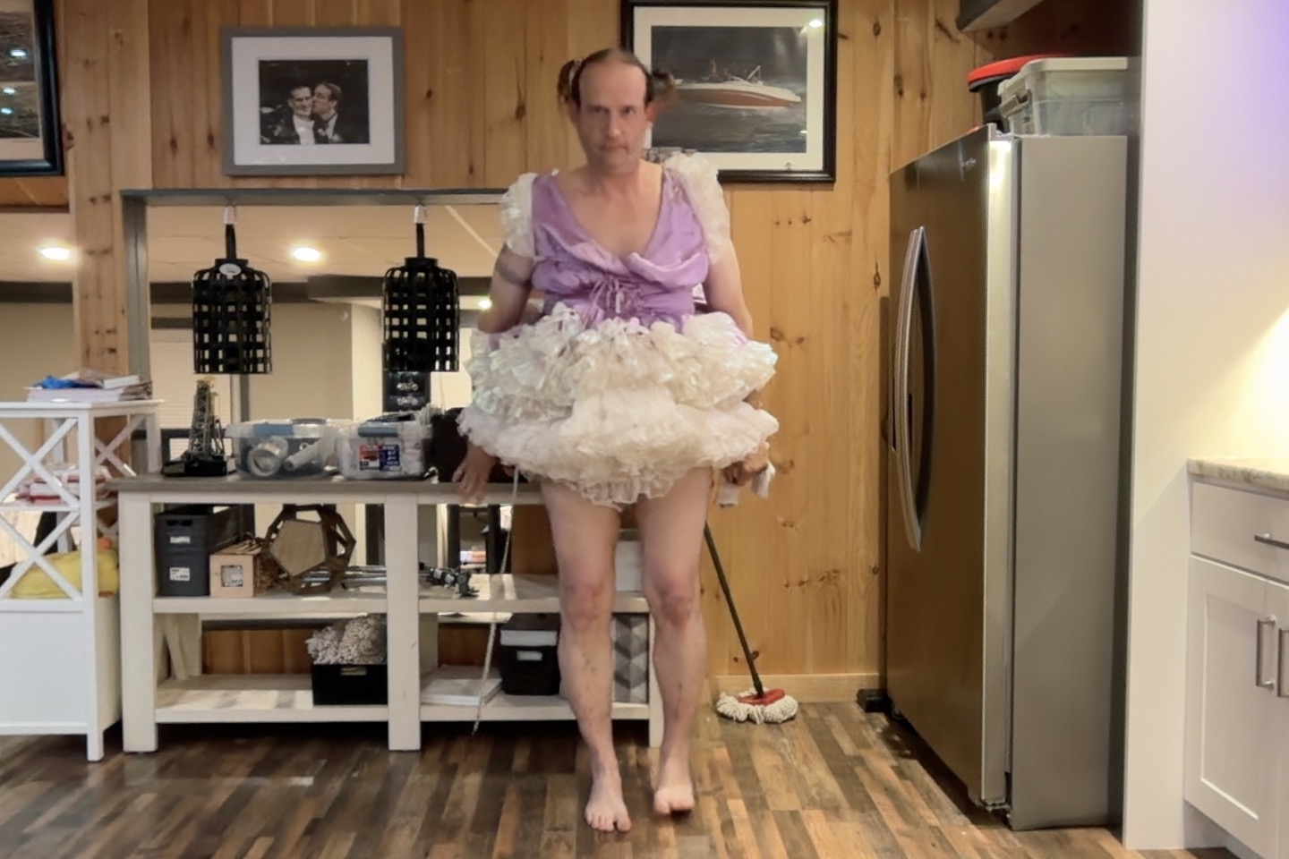 A barefoot person in a lavender top and white ruffled skirt stands in a kitchen with wood-paneled walls.