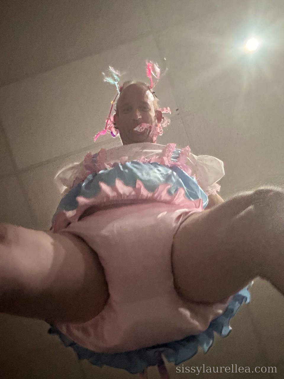 Low-angle view of a person in a pink-and-blue ruffled outfit with ribboned headpiece, shot beneath a bright ceiling light.